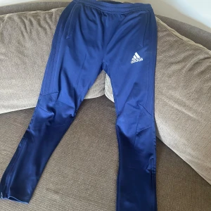 Blå träningsbyxor från Adidas - Snygga marinblå träningsbyxor från Adidas med vit logga på låret. Byxorna har smal passform, resår i midjan och dragkedjor vid bensluten. Perfekta för träning eller chill. Gjorda i glansig polyester för sportig känsla.
