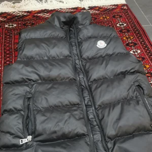 Svart dunväst från Moncler - Snygg svart dunväst från Moncler med hög krage och två dragkedjefickor. Västen har quiltad design och klassisk Moncler-logga på bröstet. Perfekt att slänga över en hoodie eller tröja för extra värme.