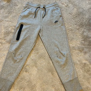 Grå mjukisbyxor från Nike med snörning - Snygga grå mjukisbyxor från Nike med svart snörning i midjan och logga på låret. Byxorna har ribbade muddar vid bensluten, dragkedjeficka på ena benet och är tillverkade i mjuk bomullsblandning. Perfekta för chill eller träning.