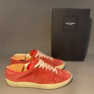 Röda mockasneakers från Saint Laurent - Snygga röda sneakers från Saint Laurent Paris i mjuk mocka med vita snören och krämfärgad sula. Låg modell med klassisk rund tå och diskreta sömmar. Perfekta för dig som vill ha en stilren och lyxig vardagssko med exklusiv känsla.