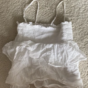 Vit volangtop från Gina Tricot - Supersöt vit topp från Gina Tricot med smockad överdel och tunna axelband. Toppen har volanger nedtill och är gjord i ett lätt och luftigt bomullstyg. Perfekt för varma dagar💕 skriv vid frågor☺️
