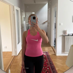 Rosa Nike Pro träningslinne - Säljer en sportig rosa Nike Pro linnetopp med vit swoosh-logga framtill. Modellen är tight och har brottarrygg för maximal rörelsefrihet. Perfekt för träning och tillverkad i ett lätt och stretchigt syntetmaterial som andas. Linnet är lite kortare än vanligt men märks knappt!!