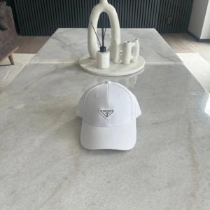 Vit keps från Prada - Snygg vit keps från Prada med klassisk böjd skärm och ikonisk metallogo framtill. Kepsen är tillverkad i bomull och har justerbar passform baktill. Perfekt accessoar för en clean och stilren look. Helt ny 