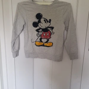 Grå Disneytröja med Mickey Mouse H&M - Superfin grå långärmad tröja från H&M x Disney med stort tryck av Mickey Mouse på framsidan. Tröjan är i mjuk bomull och har ribbade muddar vid ärmslut och nederkant. Perfekt för dig som älskar klassiska Disney-vibbar!