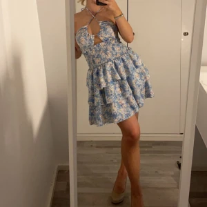 Blommig blå volangklänning - Supersöt kort klänning med blått och vitt blommönster, volanger och smockad midja. Övredelen går att justera på flera olika sätt.