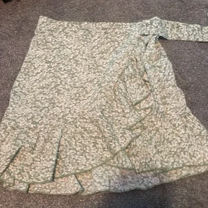 Grön blommig omlottkjol från SHEIN - Söt grön omlottkjol från SHEIN med vitt blommönster och volangkant. Kjolen har knytband i midjan och är perfekt för en somrig look. Lätt och luftig modell som ger en avslappnad vibe.