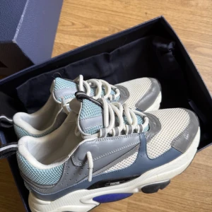 Dior B22 - Har bara använt den i 2/3 veckor säljer dem eftersom jag.. beställde fel storlek. snygga sneakers från Dior i en stilren design med vita, grå och blå detaljer. Skorna har snörning och en bekväm passform, perfekt för en trendig look. 
