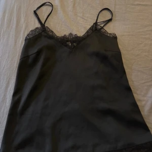 Svart slipklänning med spetsdetalj - Svart kort slipklänning med tunna axelband och v-ringning dekorerad med spets. Snygg minimalistisk look, perfekt för lager-på-lager eller som den är. Mjuk och lätt glansig yta ger en lyxig känsla.