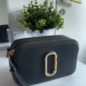 Svart axelväska från Marc Jacobs - Snygg svart axelväska från Marc Jacobs med guldiga detaljer och dubbla dragkedjor. Väskan har en stilren, boxig form och är tillverkad i strukturerat skinn. Framsidan pryds av det ikoniska metallspännet. Perfekt storlek för det viktigaste.