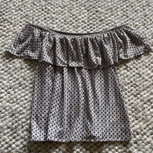 Offshoulder topp med volang från H&M - Säljer en söt offshoulder topp från H&M med volang runt hela axelpartiet. Toppen har ett småmönstrat tryck i blått och rött på vit botten och är tillverkad i ett lätt och luftigt material. Jättefin att använda nu i sommar🥰