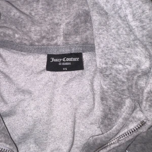 Grå hoodiejacka Juicy Couture XS - Mysig grå hoodiejacka från Juicy Couture i mjuk bomullsblandning. Jackan har huva, dragkedja framtill och två stora fickor. Perfekt för en chill och avslappnad look. Klassisk JC-logga broderad på bröstet.