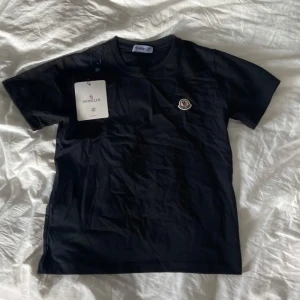 Svart Moncler t-shirt - Svart t-shirt från Moncler med klassisk rund hals och korta ärmar. På bröstet sitter Monclers ikoniska broderade logga i vitt, blått och rött. T-shirten är i mjuk bomull och har en clean, stilren look som passar perfekt till jeans eller shorts.