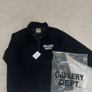Svart vindjacka från Gallery Dept. - Svart vindjacka från Gallery Dept. med vit logga tryckt på bröstet och ryggen. Jackan har dragkedja framtill och klassisk krage. Tillverkad i lätt syntetmaterial, perfekt för en clean och stilren look. Storlek M