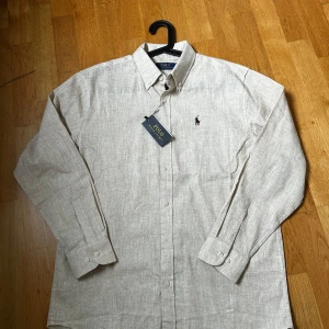 Beige linneskjorta Polo Ralph Lauren M - Snygg beige skjorta från Polo Ralph Lauren i linne med klassisk button down-krage och broderad logga på bröstet. Skjortan har lång ärm, knappar framtill och en stilren, tidlös look. Perfekt för dig som gillar en clean och avslappnad stil.