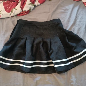 Svart kortkjol från H&M Divided - Supersöt svart kortkjol från H&M Divided med två vita ränder längst ner. Kjolen är plisserad och har en klassisk tennisstil som ger en sportig vibe. Perfekt att styla med en oversized hoodie eller crop top.