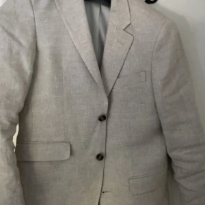 Beige linnékavaj Dressmann slim fit - Snygg beige kavaj från Dressmann i 100% linne, slim fit-modell. Klassisk design med två knappar framtill, fickor med lock och snygga detaljer vid ärmslutet. Perfekt för dig som vill ha en stilren och luftig look. Köpt för 2499kr andvänd 1 gång