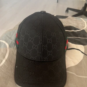 Svart Gucci keps med GG-mönster - Snygg svart keps från Gucci med klassiskt GG-monogram över hela kepsen. På sidorna finns de ikoniska gröna och röda ränderna. Kepsen har böjd skärm och är tillverkad i ett slitstarkt tyg som känns lyxigt. Och den är inte använd en ända gång