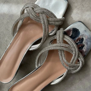 Silvriga sandaler med strass och klack - Glittriga sandaler i silver med fyrkantig tå och låg klack. Remmarna är täckta av små strasstenar i ett flätat mönster. Vet ej vilket märke de är ifrån! 