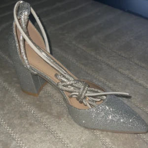 Helt oanvända Glittriga silvriga pumps med snörning - Snygga pumps i silver med glitter och spetsig tå. Skorna har en stabil blockklack och glittriga snören som går att knyta runt vristen. Perfekta för dig som vill sticka ut med extra bling. Storlek 37. Priset går att diskuteras vid snabb affär!