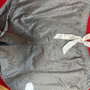 Grå mjukisbyxor med rosett XL - Grå mjukisbyxor i stretchigt material med bred resår i midjan och en snygg vit rosett framtill. Byxorna är tillverkade i en mjuk blandning av polyester och spandex, perfekta för chill dagar hemma eller när du vill vara extra bekväm.