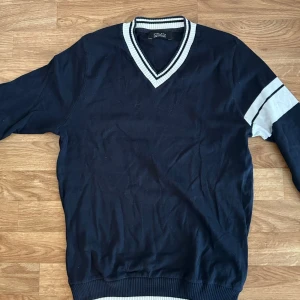 Mörkblå v-ringad tröja Zara Man - Snygg mörkblå tröja från Zara Man med v-ringad krage och vita ränder på krage, ärmslut och nederkant. Tröjan har lång ärm och sportig känsla med randig detalj på ena ärmen. Perfekt för en stilren och avslappnad look.