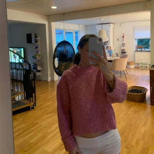 Rosa stickad tröja med hög krage - Mysig rosa stickad tröja med hög krage och breda ärmar. Tröjan har en loose passform och är croppad i modellen, perfekt att styla med höga jeans eller kjol. Ribbstickade detaljer vid krage, ärmslut och nederkant. Superfin färg som poppar!