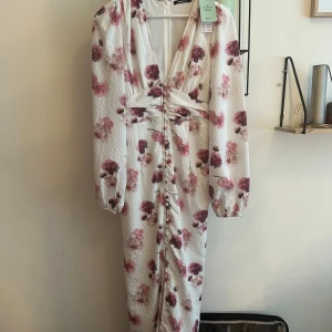Vit blommig långklänning med v-ringning - Superfin långklänning från Gina Tricot i vit bas med rosa och vinröda blommor. Klänningen har v-ringning, markerad midja, långa puffärmar och knappar framtill. Materialet är lätt strukturerat och känns luftigt. Perfekt för sommarens alla tillfällen och speciellt till midsommar. Helt ny med prislapp och slutsålt på Gina. Säljer för priset jag köpte på Plick.