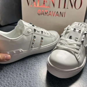 Valentino Rockstud vita sneakers - Snygga vita Valentino Rockstud sneakers i skinn med ikoniska silverfärgade nitar längs sidorna och på tungan. Klassisk rund tå, platt sula och vita snören. Perfekta för dig som vill sticka ut med en lyxig och edgy vibe.