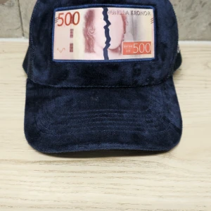 Blå keps Bruce Lee x All Cap One - Unik blå keps i samarbete mellan Bruce Lee och All Cap One. Framsidan har en patch med ett motiv av en 500-kronorssedel med konstnärlig twist. Kepsen är i mjuk manchester med mesh på sidorna och broderad logga på sidan.