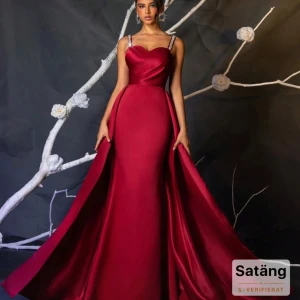 Vinröd långklänning med smala axelband - Magisk vinröd långklänning i satin med figurnära passform och bred, svepande kjol. Klänningen har smala axelband med glittrande detaljer och en snygg sweetheart-ringning. Perfekt för dig som vill sticka ut på bal eller fest.