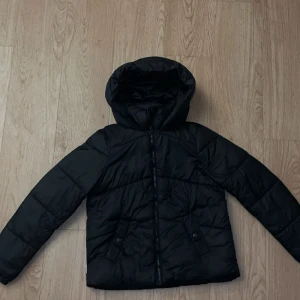 Svart pufferjacka från Vero Moda XS - Svart pufferjacka från Vero Moda i storlek XS. Jackan har huva, dragkedja framtill och två fickor med knappstängning. Perfekt för kalla dagar och enkel att matcha med allt. Snygg och stilren look som funkar till alla outfits.