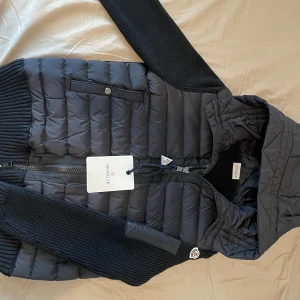 Moncler cardigan i nyskick - Exklusiv svart Moncler med quiltad kropp och stickade ärmar. Huva, dragkedja, knappfickor och klassiska loggan på ärmen. Äkthetsintyg med både NFC och QR-kod som leder direkt till Monclers officiella hemsida. Perfekt mix av stil och värme – både street och lyx i ett.  Pris kan diskuteras – kom med förslag. Först till kvarn.