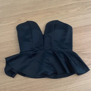 Svart peplum korsett-topp - Supersnygg svart korsett-topp med peplumvolang och djup v-ringning. Axelbandslös modell som sitter tight och ger en riktigt cool siluett. Perfekt att styla med jeans eller kjol för en trendig look.