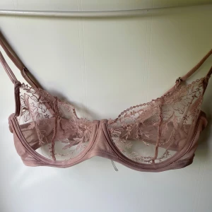 Rosa spets bralette från Ella M. - Superfin ljusrosa bralette från Ella M. med tunna axelband och transparent mesh i spets med broderade blommor. Snygg och feminin design med mjuka kanter och justerbara band. Perfekt att styla under en öppen skjorta eller kavaj.