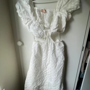 Vit broderad klänning Love Triangle - Superfin vit klänning från Love Triangle med broderade blommor, puffärmar och snygg cut-out i midjan. Klänningen har knytning framtill och är perfekt för sommarens alla tillfällen. Lätt och luftig med romantisk känsla.