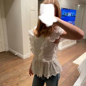 Vit blus med volang och broderi - Superfin vit blus med broderade blommor och volangdetaljer längs axlar och bröst. Blusen har rund hals, korta ärmar och en lätt utsvängd nederdel. Perfekt för dig som gillar romantisk och söt stil. Dragkedja i ryggen för enkel påklädning.