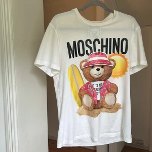 Vit Moschino t-shirt med björntryck - Snygg vit t-shirt från Moschino med stort tryck av en nalle i semesterlook, surfbräda och sol. T-shirten har rund hals och korta ärmar. Perfekt statement-plagg för dig som gillar lekfull design och coola prints.