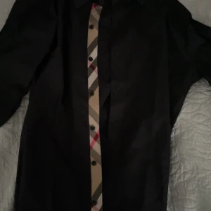 Svart Burberry skjorta med rutiga detaljer - Svart skjorta från Burberry med klassisk krage och långa ärmar. Framsidan har Burberrys ikoniska beige, svarta och röda rutiga knappslå. Broderad TB-logga på bröstet. Skjortan är i  bummul och siden och har en clean, stilren look som sticker ut med de rutiga detaljerna. Helt ny med tags fortfarande kvar 