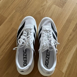 Adidas Adizero vita löparskor - Endast använt två gånger. Snygga Adidas Adizero sneakers i vitt med svarta ränder. Lätta och ventilerande med textilöverdel, snörning och platt sula. Perfekta för löpning eller träning, med Continental-gummisula för extra grepp. Stilren och sportig design.