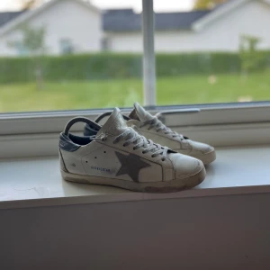 Golden Goose  - Golden Goose sneakers i vitt skinn med grå mockadetaljer och ikonisk stjärna på sidan. Skorna har grå snörning, mörkblå häl och platt sula i gummi. Perfekta för en avslappnad och trendig look. 