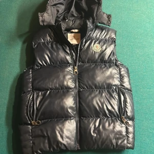 Svart dunväst från Moncler - Säljer en snygg svart dunväst från Moncler med dragkedja och huva. Västen har två praktiska fickor med dragkedjor och Monclers logga broderad på bröstet. Perfekt för kyligare dagar. Skicket är 8/10