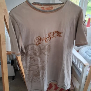 Beige t-shirt från Quiksilver med tryck - Säljer en beige t-shirt från Quiksilver med coolt tryck och orange text framtill. T-shirten har korta ärmar, rund hals och kontrastsömmar vid ärmslut och hals. Ficka på vänster arm. Cool surfer vibe till sommaren 😎