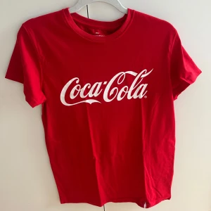 Röd Coca-Cola t-shirt - Röd t-shirt från Coca-Cola med klassisk vit logga framtill och liten vit flaska bak i nacken. Skön passform, rund hals och korta ärmar. Per för dig som gillar retro vibes och ikoniska prints