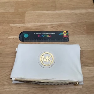 Vit kuvertväska Michael Kors skinn - Snygg vit kuvertväska från Michael Kors i skinn med guldfärgade detaljer och dragkedja. Väskan har ett stort MK-emblem framtill och magnetknappar. Insidan är fodrad med logomönstrat tyg. Perfekt för att bära det viktigaste med stil.