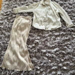 Skjorta och kjol - Säljer en beige långkjol i storlek S tillsammans med en cremfärgad skjorta storlek S. Kjolen och skjortan har ett vågigt, ribbat mönster och är gjord i ett mjukt, lätt material som ger en flowy känsla. Perfekt för dig som gillar stilrena och trendiga plagg med en twist.