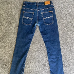 Nudie Jeans Thin Finn blå jeans 33/32 - Säljer ett par Nudie Jeans Thin Finn i klassisk blå tvätt. Jeansen är tillverkade av 98% ekologisk bomull och 2% elastan för extra stretch. De har smal passform, snygga bakfickor med vågig söm och orangea detaljer. Perfekta för dig som gillar stilrena och bekväma jeans.