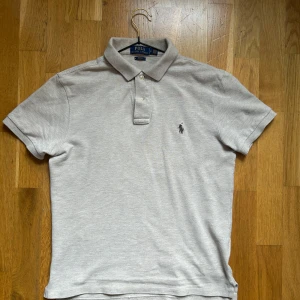 Beige pikétröja från Polo Ralph Lauren - Snygg beige pikétröja från Polo Ralph Lauren i 100% bomull. Klassisk krage, två knappar och den ikoniska broderade loggan på bröstet.