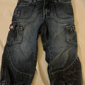 Blå cargopants från Detroit, stl 86( barn st) - Säljer ett par mörkblå cargopants från Detroit i jeansmaterial. Byxorna har flera fickor, rosa dekorativa knappar och sömmar samt broderade detaljer på benen. Perfekta för dig som gillar en cool och avslappnad stil.