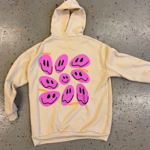 Beige hoodie med rosa smileys - Beige hoodie med huva och snörning. Baksidan har stora rosa smiley-ansikten och texten 'HAPPY' i orange. Mjuk och skön, perfekt för dig som gillar färgglada och unika plagg. Långärmad och loose fit för en avslappnad stil.