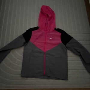 Nike  - Nike vindjacka med snygg färgblock-design i rosa, grått och svart. Jackan har huva, hel dragkedja och lång ärm. Perfekt för blåsiga dagar och sportiga outfits. Ikonisk Nike-logga på bröstet för extra stilpoäng.